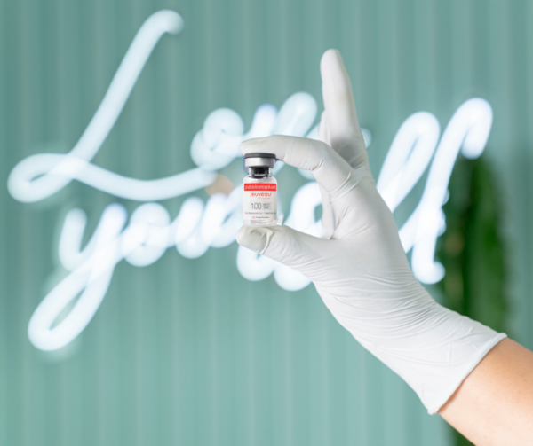 Gloved hand holding a small vial of botulinum toxin in front of a neon Love Yourself sign