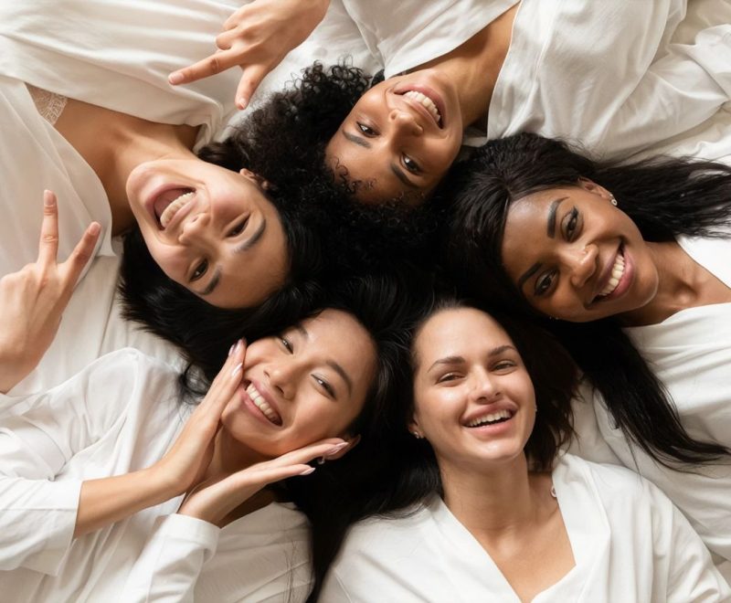 Five smiling women in white robes lying in a circle with their heads together